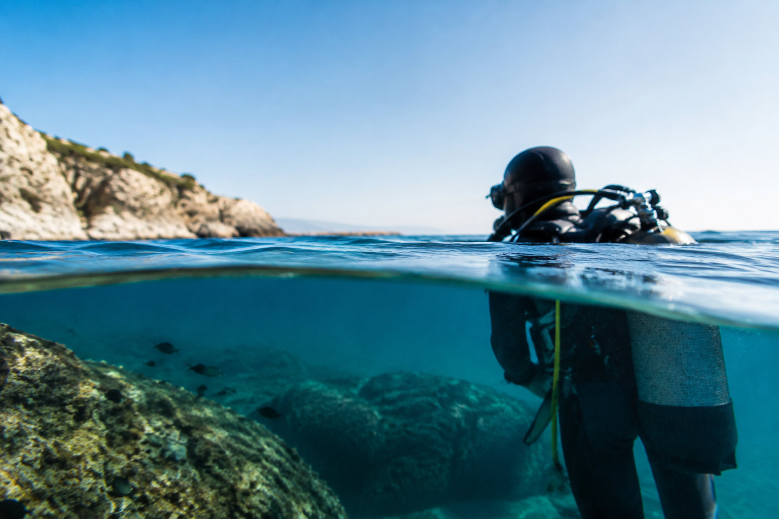 Un plongeur sous-marin avec équipement vu à la surface de l'eau, près d'une côte rocheuse sous un ciel bleu.